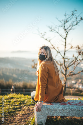 Woman on a yellow coat sitting on a bench with a face mask