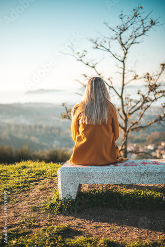 Slika na platnu Woman on a yellow coat sitting on a bench contemplating the landscape