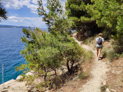 Young active feamle tourist wearing small backpack walking on coastal path among pine trees looking for remote cove to swim alone in peace on seaside in Croatia. Travel and adventure concept.