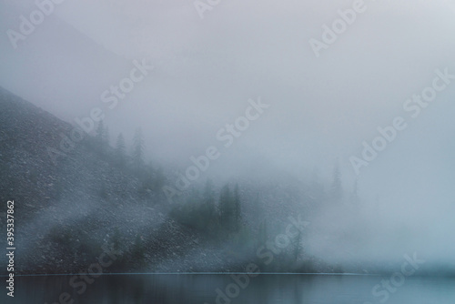 Dense fog above tranquil mountain lake. Calm water and stony steep slope with coniferous trees among low clouds. Ghostly atmospheric view. Minimalist scenery in mysterious place at early foggy morning