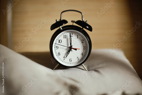 Classic black alarm clock showing seven o'clock with a bed in the background, waking up in the morning concept