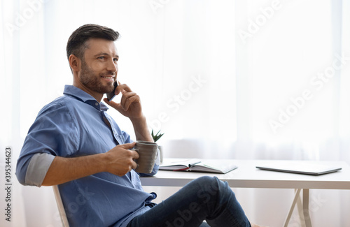 Handsome freelancer man talking on cellphone and drinking coffee at home
