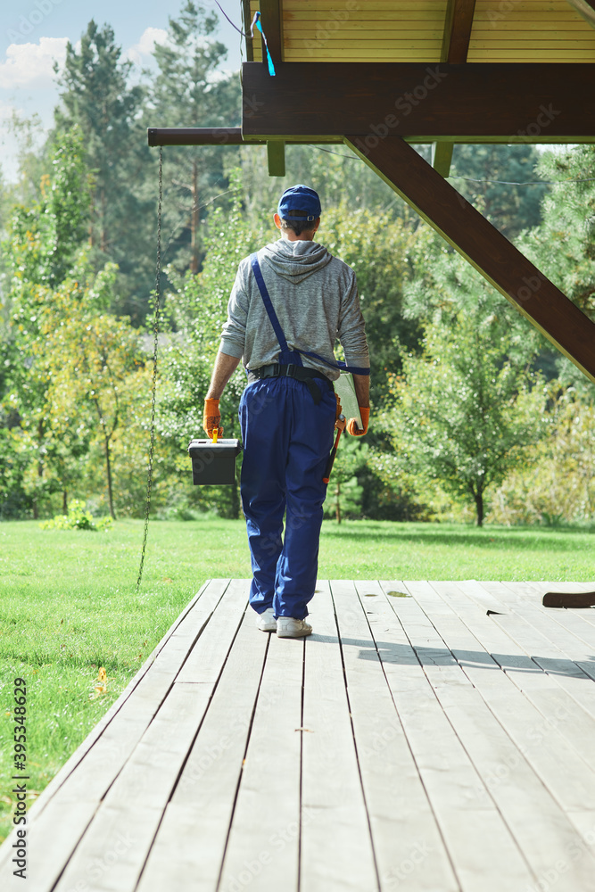 Full length shot of young workman in blue overalls carrying toolbox after doing repair work in the cottage house on a sunny summer day