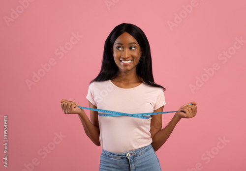 Weight loss and healthy diet concept. Happy African American lady measuring her breasts on pink studio background