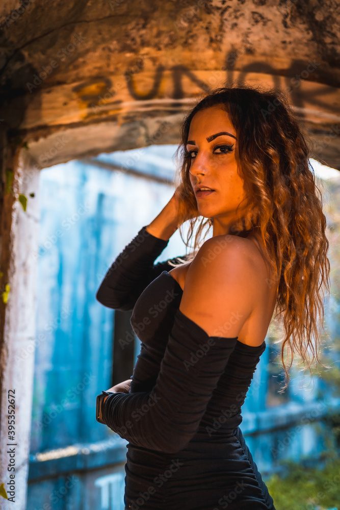 Obraz premium Lifestyle, Caucasian girl with wavy hair, in a tight black dress and heels inside a vandalized building. Portrait in window of building interior