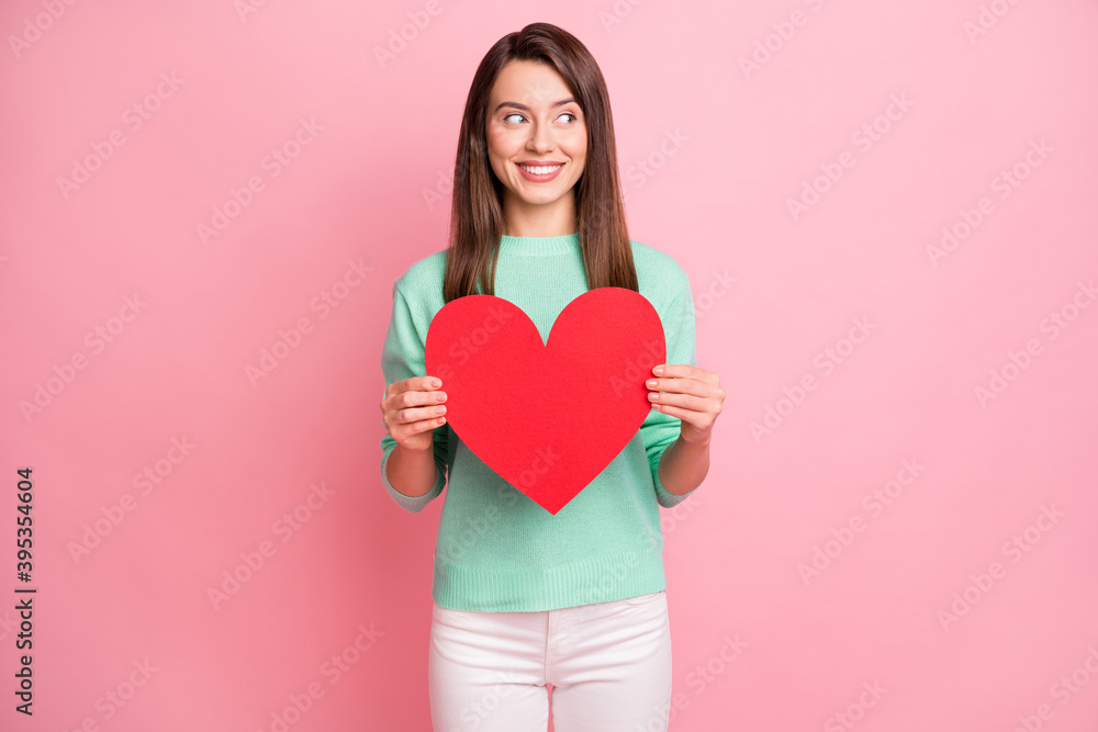 Portrait of attractive cheerful girl holding in hands red heart looking aside isolated over pink pastel color background Portrait of attractive cheerful girl holding in hands red heart looking aside isolated over pink pastel color background