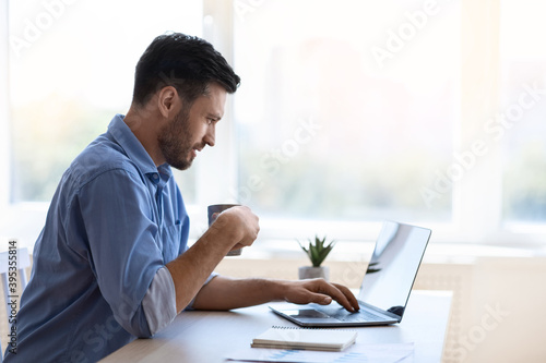 Handsome male freelancer browsing social media on laptop and drinking morning coffee