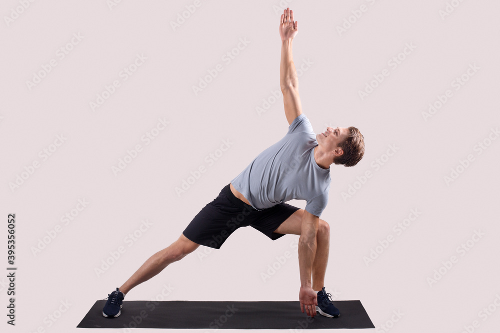 Strong young man standing in yoga pose on sports mat over light studio background, full length portrait
