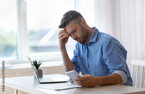 Worried male freelancer looking at smartphone screen while working at home office photo
