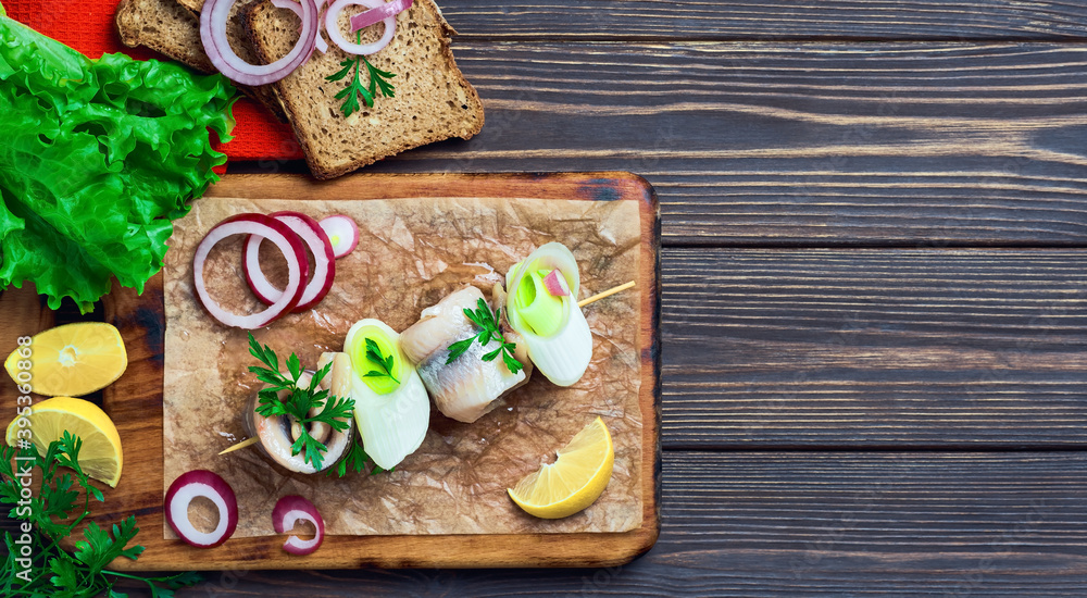 Herring roll on a skewer with parsley, onion and lemon wedge, top view. The sandwiches are arranged on a cutting board, on a dark wooden background with space for text. Fast food or snack