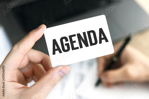 Text AGENDA on card in hand on the background of laptop and documents.