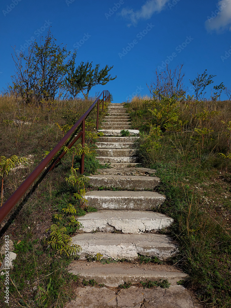 Old concrete stairs with rusty iron pipe handle leading to the blue sky. High hill with trees and green grass. Beautiful landsaft. Rural style.