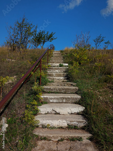 Old concrete stairs with rusty iron pipe handle leading to the blue sky. High hill with trees and green grass. Beautiful landsaft. Rural style.