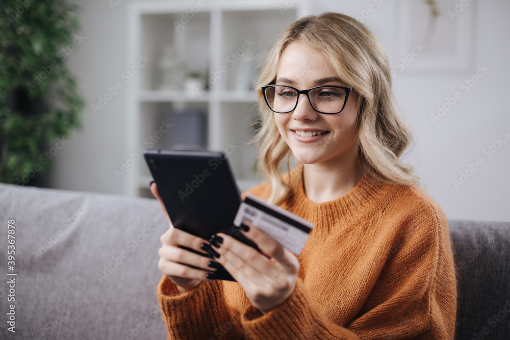 Happy young woman in eyeglasses using digital tablet and credit card for online shopping while staying at home. Concept of modern technology. Happy young woman in eyeglasses using digital tablet and credit card for online shopping while staying at home. Concept of modern technology.