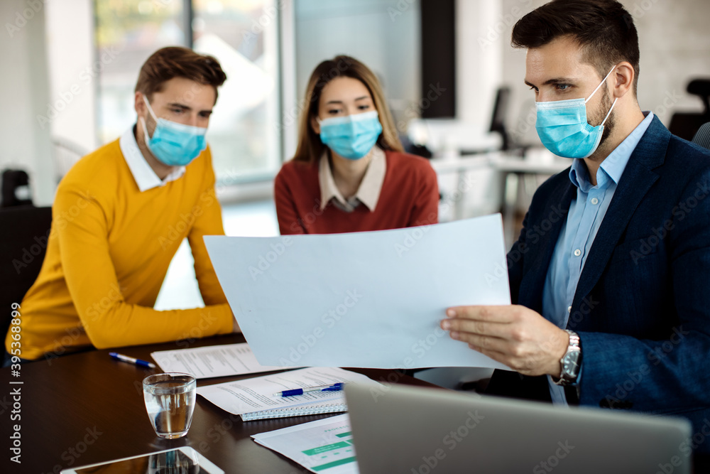 Real estate agent and a couple analyzing housing plans on a meeting while wearing protective face masks.