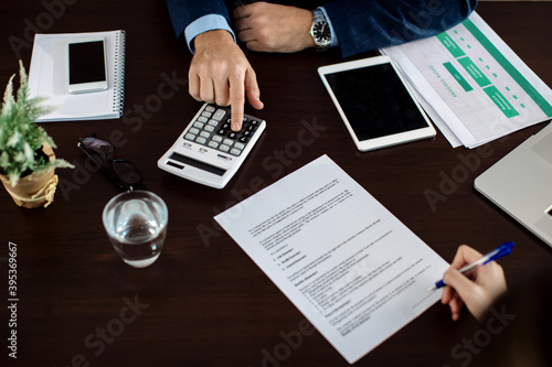 Close-up of bank manager calculating costs while clients is singing a contract in the office.