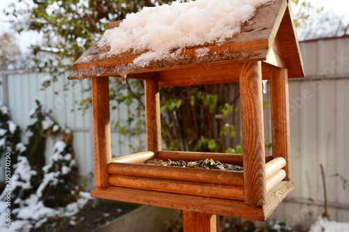 Canvas Print A close-up of an installed on a wooden pole, post solid wood chalet-style bird feeder with sunflower seeds inside to feed hungry wild birds at home during snowy winter