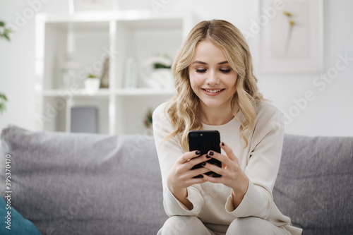 Smiling young lady with wavy blond hair using modern smartphone while sitting on grey couch. Concept of people, technology and leisure time.