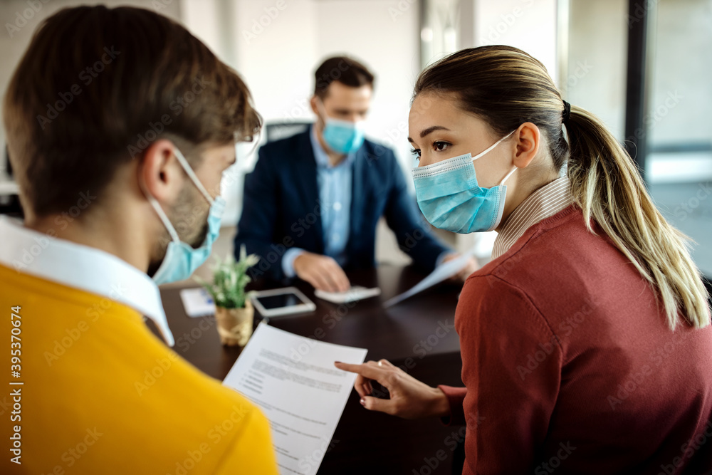 Young couple with face masks discussing about paperwork during a meeting with their financial advisor. Young couple with face masks discussing about paperwork during a meeting with their financial advisor.