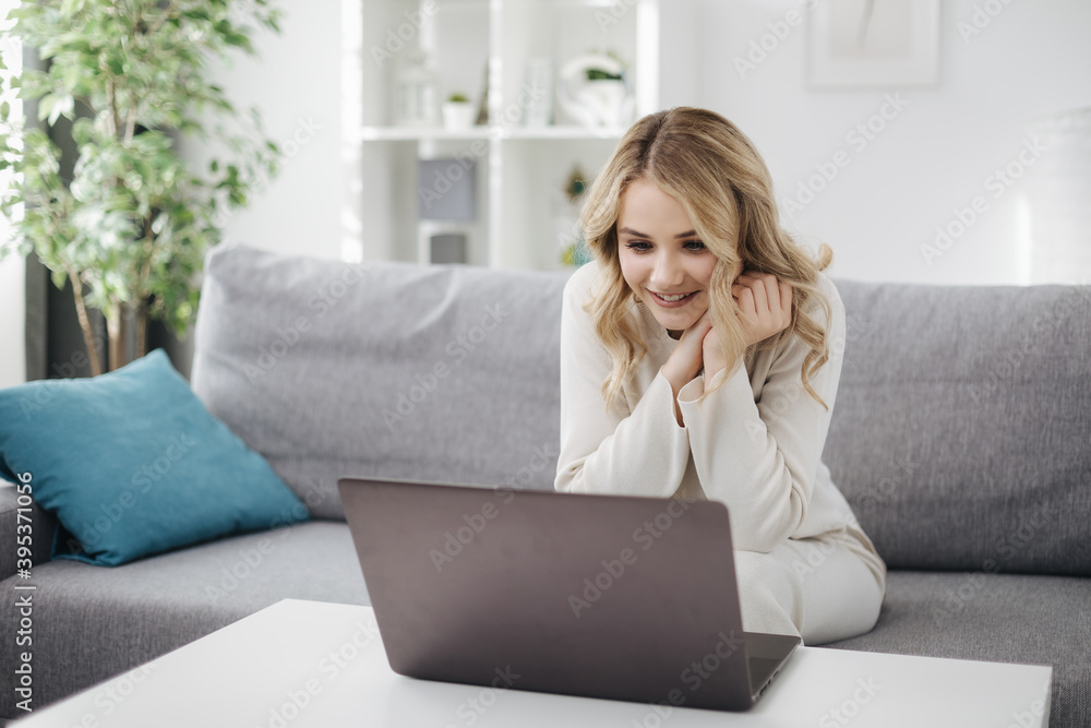 Happy young blonde in domestic outfit sitting on couch and having video conference on wireless laptop. Online conversation and modern technology concept. Happy young blonde in domestic outfit sitting on couch and having video conference on wireless laptop. Online conversation and modern technology concept.
