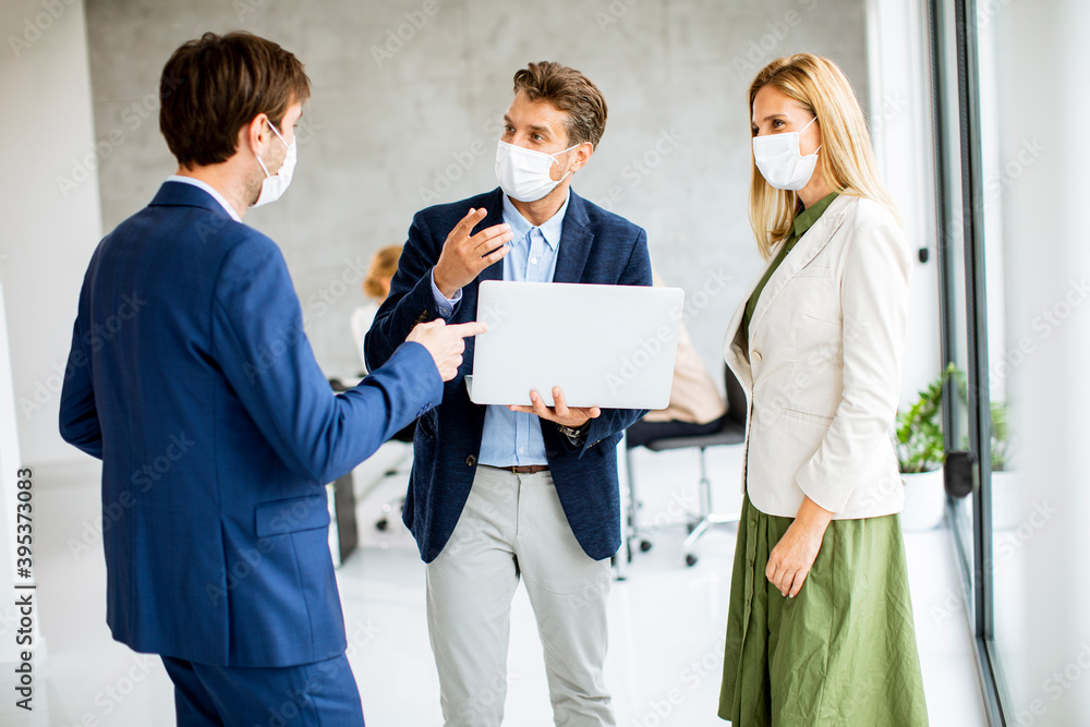 Two men and woman standing with laptop in hands indoors in the office with young people works behind them Two men and woman standing with laptop in hands indoors in the office with young people works behind them