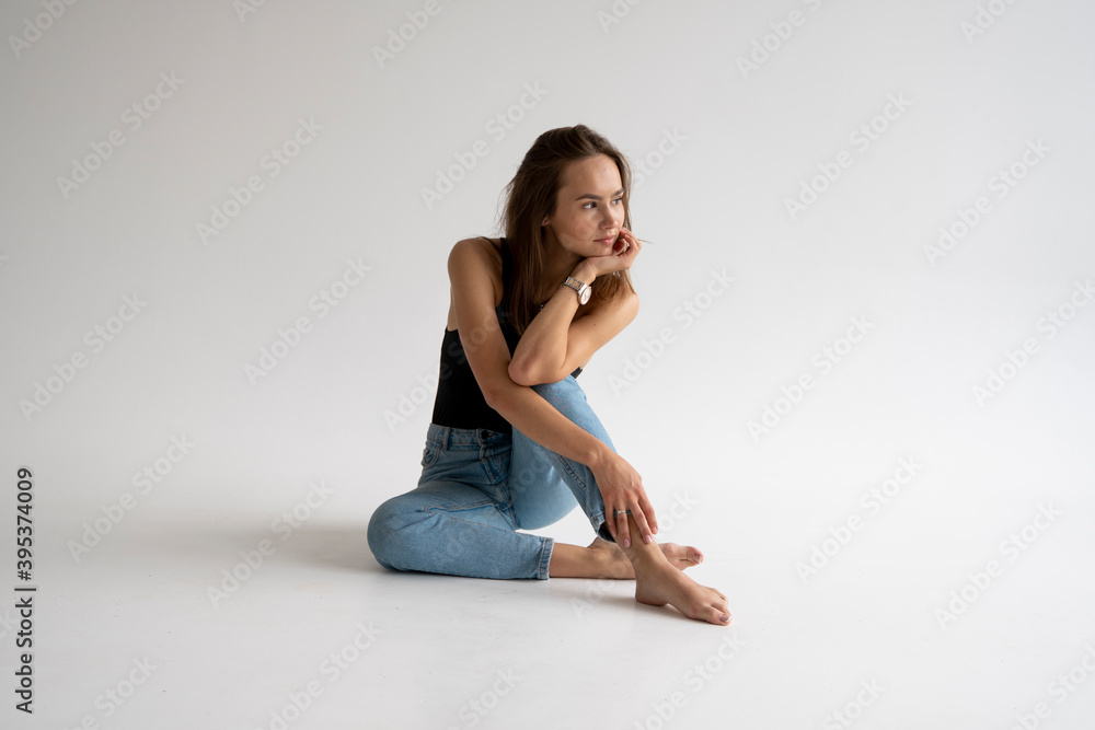 Portrait of happy young pensive woman posing in black underwear and blue jeans without a shoes, sitting on a white floor in white studio. Model tests of pretty girl in basic clothes on cyclorama. Portrait of happy young pensive woman posing in black underwear and blue jeans without a shoes, sitting on a white floor in white studio. Model tests of pretty girl in basic clothes on cyclorama.