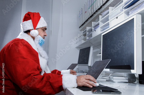 Babbo Natale con mascherina chirurgica abbassata , seduto in ufficio utilizza il computer per fare degli ordini via internet photo