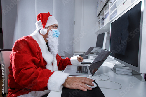 Babbo Natale con mascherina chirurgica, seduto in ufficio utilizza il computer per fare degli ordini via internet photo