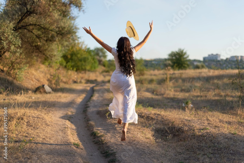 young girl in a white dress jumping in the field, tossing her hat up. Happy girl on a summer evening, against the backdrop of the setting sun