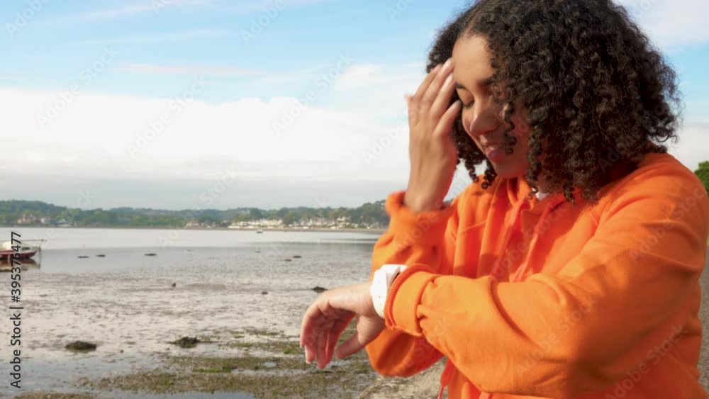 Beautiful mixed race African American girl teenager young woman wearing orange hoodie, sitting on a sea front using her smart watch for social media, communications or text messaging Beautiful mixed race African American girl teenager young woman wearing orange hoodie, sitting on a sea front using her smart watch for social media, communications or text messaging