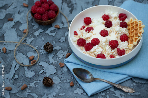 Cottage cheese in a plate with yoghurt, sprinkled with raspberries and muesli, blue on a towel, metal spoon and nuts.