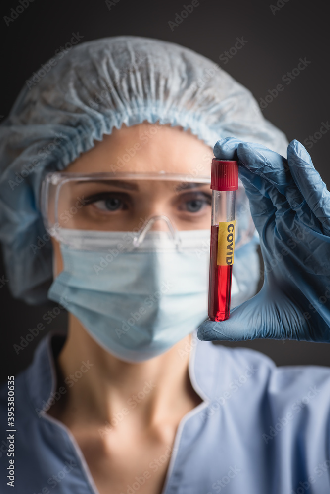 test tube with covid lettering on hand of nurse in medical cap, mask and goggles on blurred isolated on dark grey