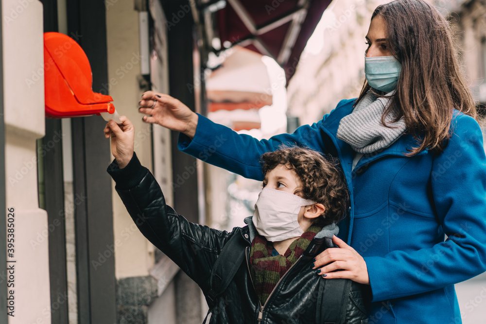 Fototapeta premium Mother and son getting the queue ticket at a local shop because of the restrictions of the 2019 coronavirus pandemic. The kid and his mom are wearing the face mask. COVID-19 lifestyle concept.