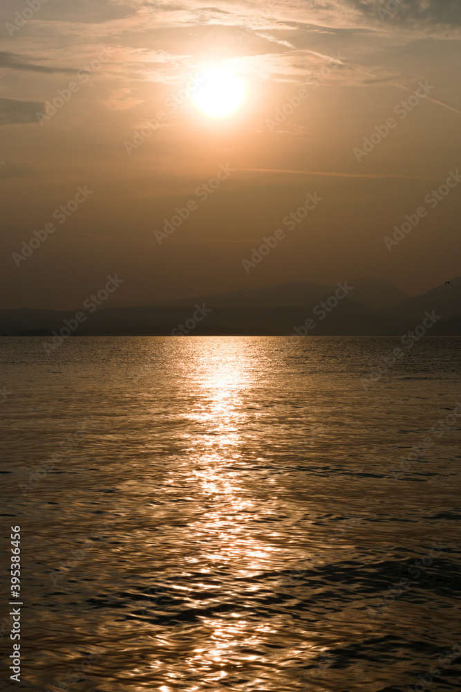 Naklejka premium Sunset at Garda lake, Italy. Sun behind clouds with reflections. 