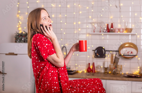 Happy laughing and smiling young beautiful woman speaking or talking by mobile phone in kitchen in red pygamas holding tea mug. New year and xmas alone online at home photo