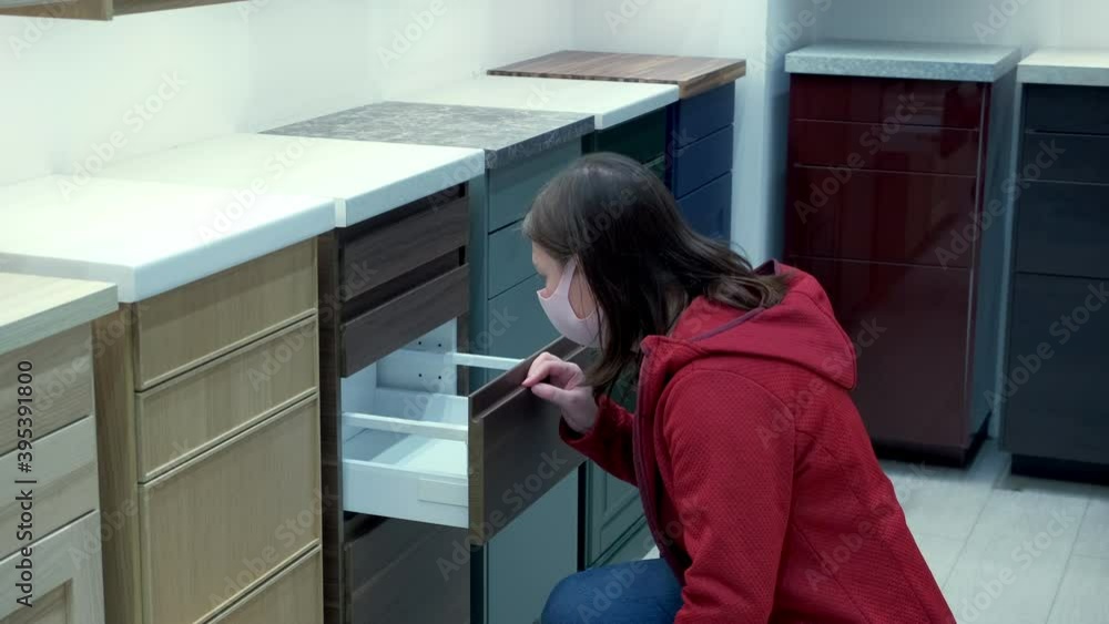 Caucasian woman in a pink, protective mask chooses the kitchen in the store. Opens the desk drawers and looks in them. Woman is wearing a red jacket Caucasian woman in a pink, protective mask chooses the kitchen in the store. Opens the desk drawers and looks in them. Woman is wearing a red jacket
