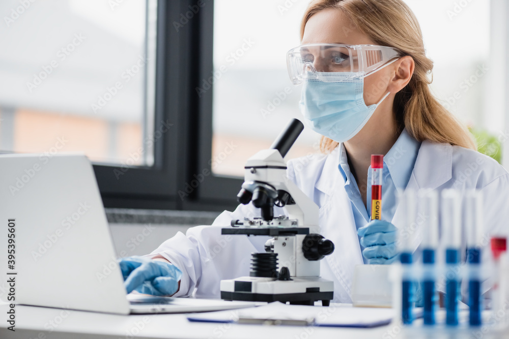 scientist in medical mask and goggles holding test tube with covid lettering and using laptop, stock image scientist in medical mask and goggles holding test tube with covid lettering and using laptop, stock image
