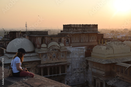 Wall Mural Young woman and tourist, sitting on the rooftop and is watching the sunset over the city skyline with a camera around her neck