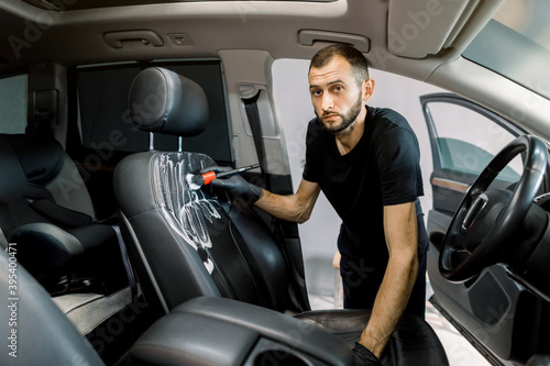 Car detailing and cleaning concept. Young man, car wash worker, wearing protective rubber gloves, cleaning the car seat of the modern vehicle, using special brush and foam, looking at camera © sofiko14
