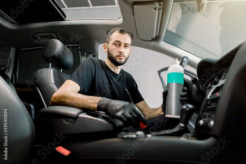 Young concentrated male car service worker cleaning dust from interior of luxury car, using special brush and plastic care professional product in bottle. Car detailing, interior care. Focus on face