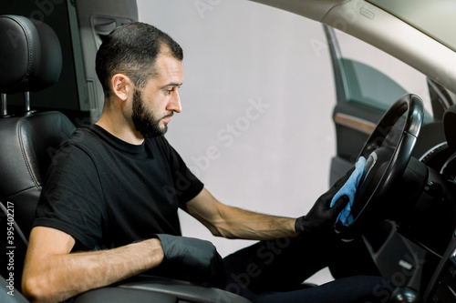 Handsome Caucasian young man in protective gloves cleaning car interior, car steering wheel using microfiber clothes. Car detailing or valeting concept. Selective focus © sofiko14