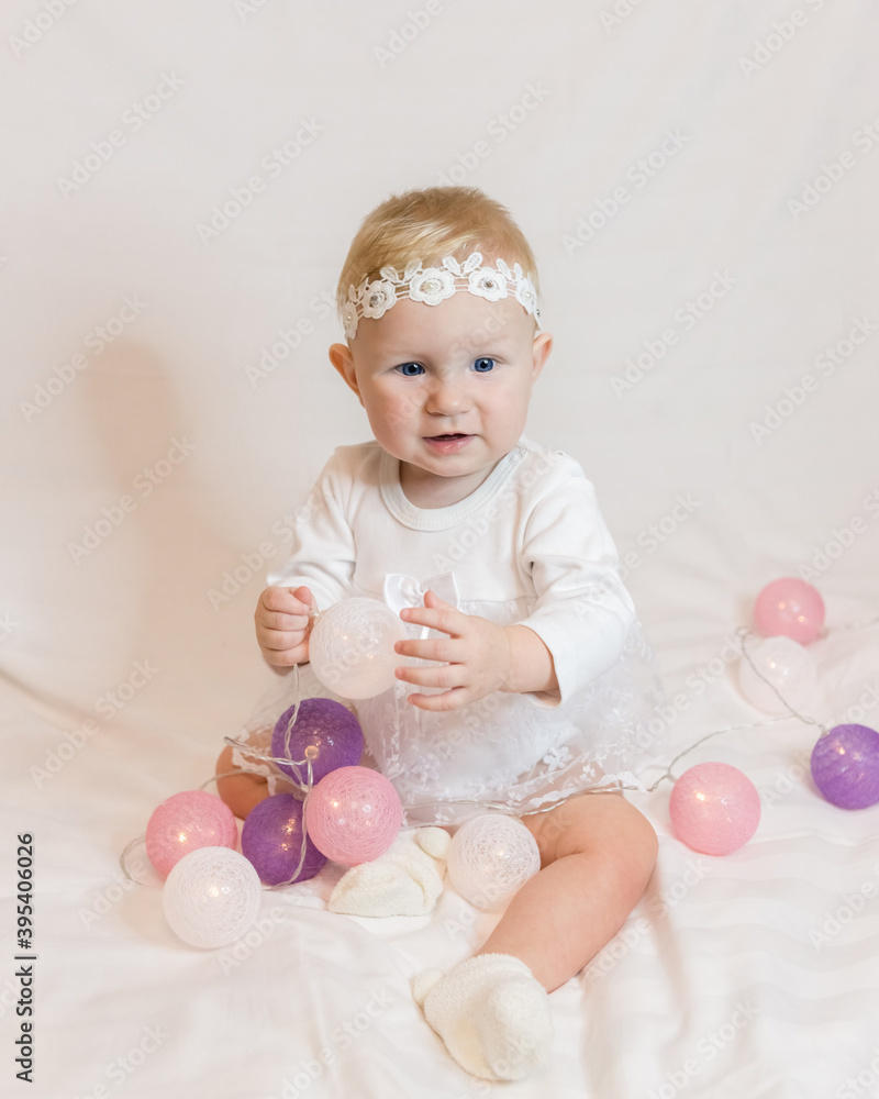 A small eight-month-old girl in a smart white dress with garland balloons in her hands is smiling. On the head of Lacy white bandage A small eight-month-old girl in a smart white dress with garland balloons in her hands is smiling. On the head of Lacy white bandage