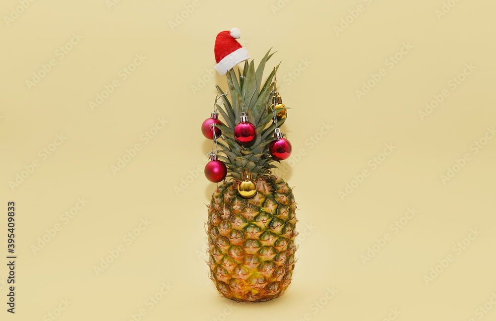 Ripe beautiful pineapple decorated with Christmas balls and Santa hat on a light yellow background. Creative idea of a Christmas tree for vegetarians.