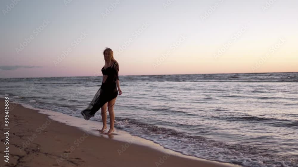 Pretty young blonde woman walks at you along sea coast during sunset at black lingerie