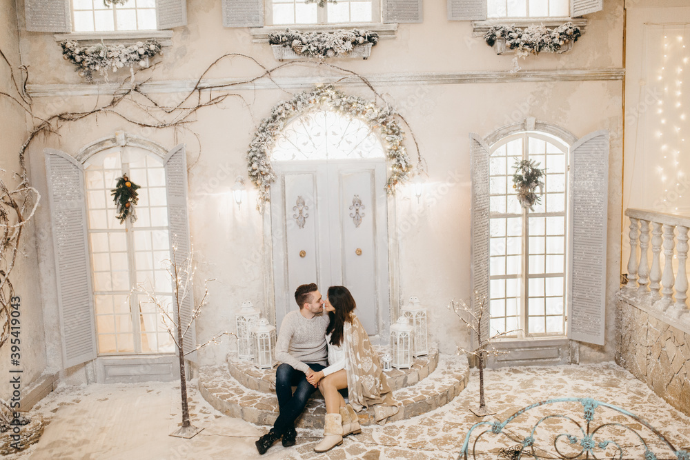 Adorable couple sit on stairs at the background Christmas decoration, cuddling, beautiful wife with loving husband spend winter holidays together, New Year celebration concept