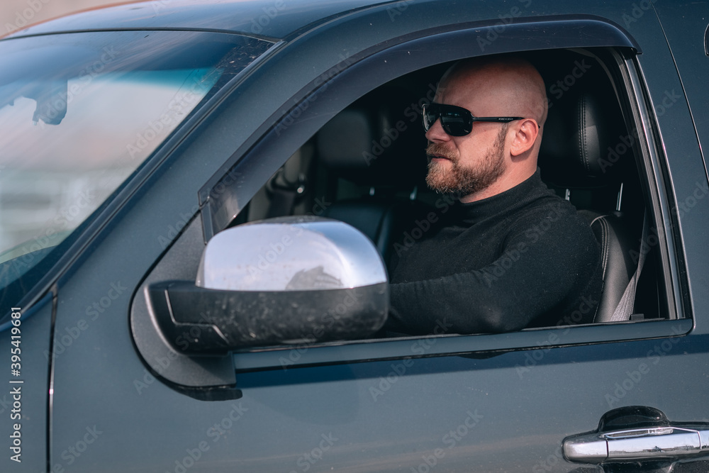 bald and bearded caucasian man in glasses with a clock in a suit behind the wheel of a black car