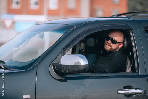 bald and bearded man in glasses with a clock in a suit behind the wheel of a black car