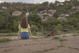 A shot of a girl sitting alone on the platform of an old station and looking at the city. From a high point overlooking a small town; next to the girl her bike