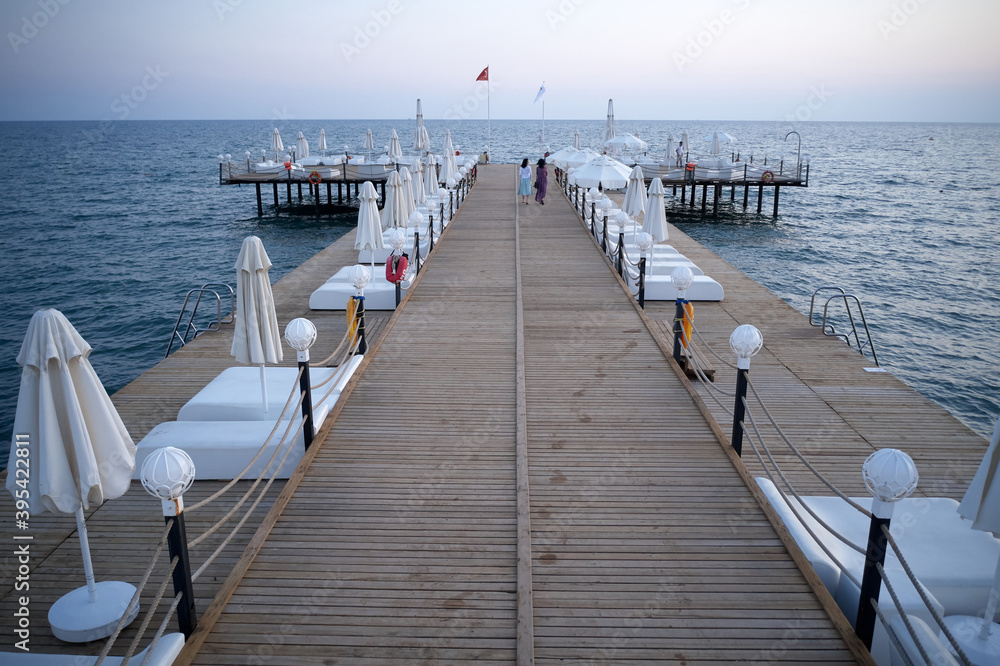 Wooden bridge with sunbeds in the sea at sunset. Beautiful landscape of calm ocean waves and long wooden pier with walking tourists. Travel to Turkey.