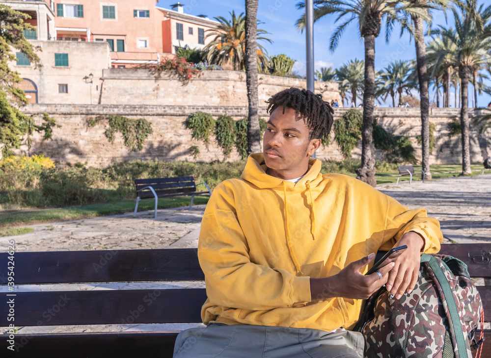 Fototapeta premium Afro american black man with yellow sweatshirt looking at smartphone laying on a wooden bench in a park